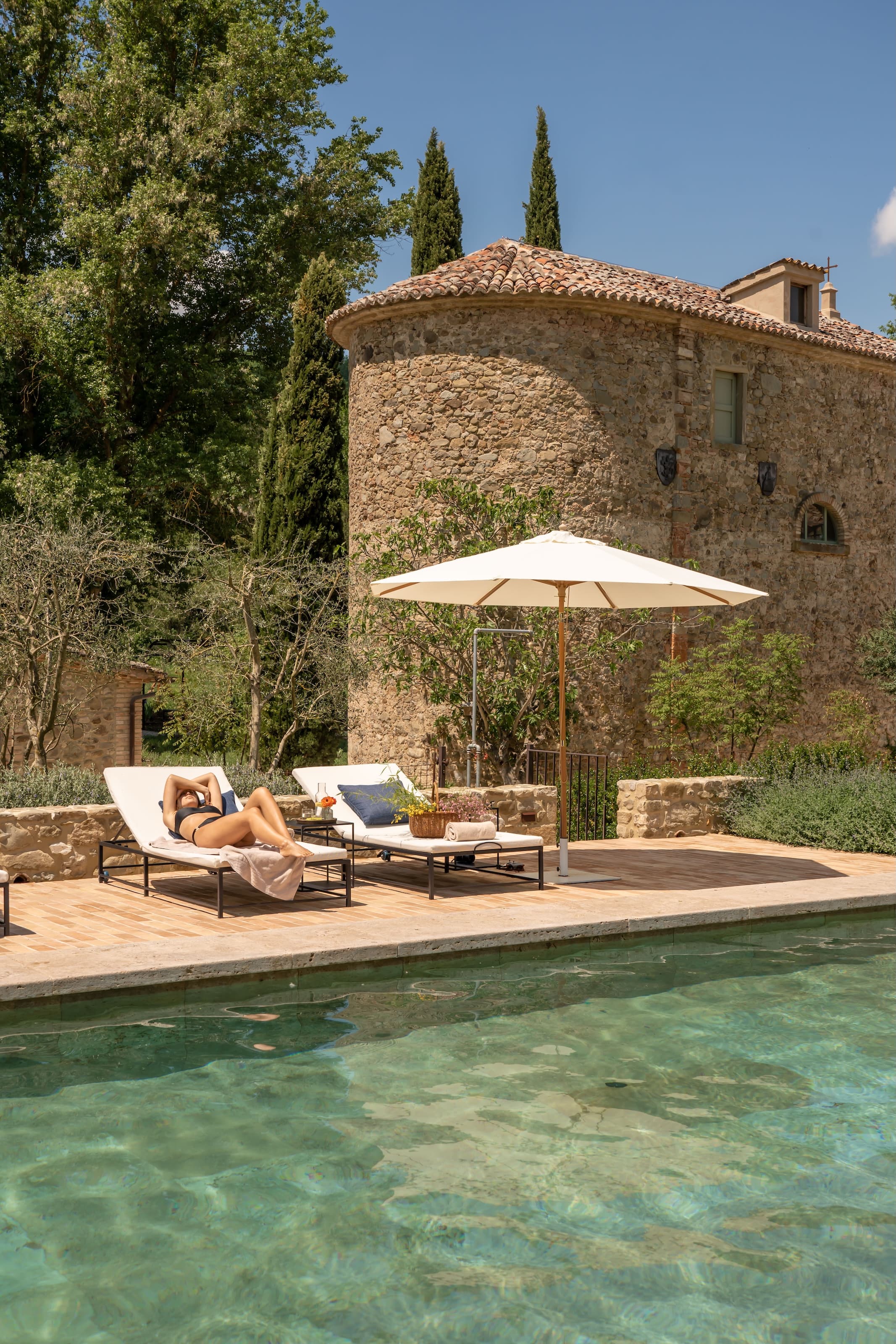 Woman sunbathing on a lounger beside the pool of a rustic stone villa, surrounded by trees and cypress trees.